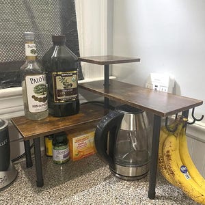 May include: A three-tiered, brown and black kitchen shelf unit with a stainless steel electric kettle, bananas, and various bottles and containers. The shelf has a rustic, industrial design.