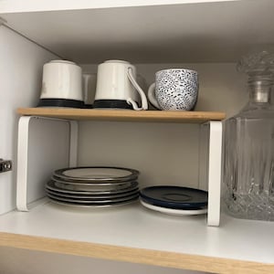 May include: A white cabinet interior with a wooden shelf holding two white mugs with black bases and a blue and white patterned mug. Below, a stack of plates with silver rims and a blue plate.