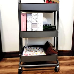 May include: A gray metal rolling cart with three shelves. The top shelf holds a small box and a plaid container. The middle shelf has recipe books and the bottom shelf has a recipe book and a wooden box. The cart has wheels.