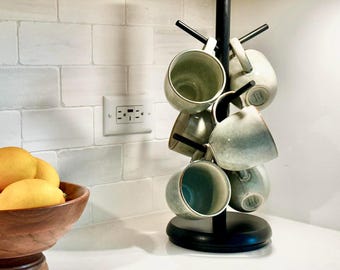 May include: A black metal mug tree holds several gray-green ceramic mugs. The mugs are arranged around the central pole. A wooden bowl filled with lemons sits in the foreground. The background features a white brick-patterned wall.