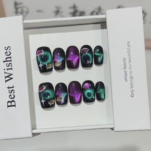 Puede incluir: Un juego de diez uñas postizas negras con un diseño temático espacial. Cada uña presenta un fondo oscuro con colores verdes, morados y azules arremolinados, y un pequeño planeta y estrellas. Las uñas se presentan en una caja blanca con el texto "Best Wishes".