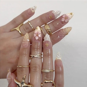 Puede incluir: Una mano con uñas acrílicas largas en forma de almendra. Las uñas presentan una variedad de diseños, incluyendo flores 3D, acentos de conchas marinas y adornos dorados. Varios anillos de oro adornan los dedos, agregando a la estética elegante. Las uñas están pintadas en una paleta de colores suaves y naturales.