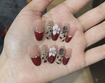 Può includere: Unghie finte con un design a manicure francese. Le unghie presentano una base nude e una punta rosso scuro, decorate con strass, decorazioni floreali e glitter. Le unghie di forma ovale sono esposte su una mano.