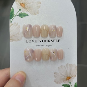 Può includere: Due set di unghie finte con un design marmorizzato e dettagli dorati. Le unghie sono nelle tonalità del rosa, beige e oro. Il testo "LOVE YOURSELF" e "To the best of you" è stampato sulla carta.
