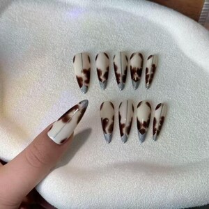 以下が含まれることがあります： 牛柄のデザインの人工爪10本セット。爪はアーモンド型で、白いベースに茶色の斑点があります。各爪には銀色の先端が付いています。