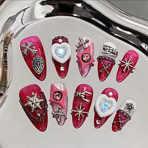 May include: A collection of ten long, almond-shaped artificial nails. Each nail is painted a deep pink or light pink and decorated with silver charms, including crosses, stars, hearts, and spiderwebs. Some nails have rhinestones.