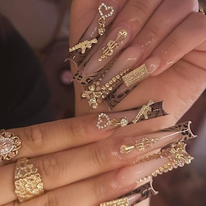 May include: Long, almond-shaped acrylic nails with a nude base and leopard print accents. The nails are decorated with gold charms, including guns, dollar signs, hearts, and bows, for a glamorous and edgy style.