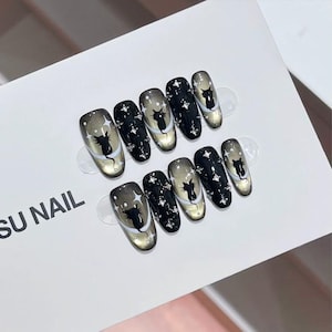 May include: A set of ten oval false nails. Each nail has a black cat silhouette, silver star accents, and a gradient design of black, silver, and gold. The nails are displayed on a white card with the text "SU NAIL".