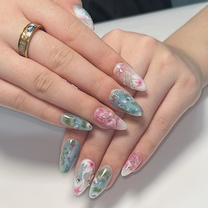 Puede incluir: Manos con uñas largas en forma de almendra con diseños florales en tonos rosa, azul y verde. Algunas uñas tienen adornos plateados. También se ve un anillo de oro con una incrustación decorativa.