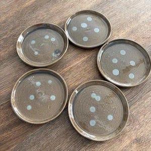 Könnte beinhalten: Fünf handgefertigte Keramikplatten mit brauner Glasur und hellblauen Tupfen. Die Teller sind rund mit leicht erhöhtem Rand. Sie liegen auf einer Holzoberfläche. Die Teller sind neutralfarben und eignen sich zum Servieren von Speisen.