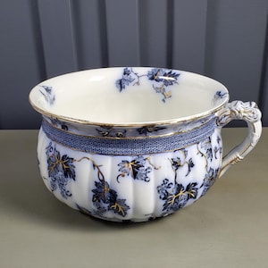 Op de afbeelding: Antieke witte porseleinen po met blauwe bloemmotieven en gouden details. De pot heeft een decoratieve handgreep en een blauw patroonband. De rand is afgewerkt met goud.