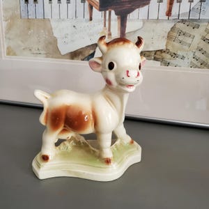May include: A ceramic figurine of a calf, featuring a cream-colored body with brown patches and horns. The calf stands on a green base, with a smiling expression and pink cheeks. The figurine is a decorative item.