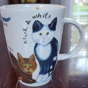 May include: A white ceramic mug featuring a hand-painted illustration of a black and white cat and a brown cat. The text "Black & white" is written in black script above the black and white cat. The mug has a curved handle and a smooth finish.