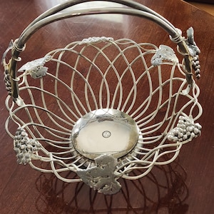 May include: Silver-toned metal fruit basket with a handle. The basket features a woven design and is adorned with grape and leaf accents. The base is a solid, reflective silver disc. A decorative piece for the home.