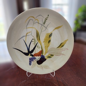 May include: A cream-colored ceramic plate with a hand-painted design of a swallow, wheat, and other foliage. The swallow is black with long, pointed wings. The wheat is yellow and green, and the foliage includes orange, purple, and white elements.