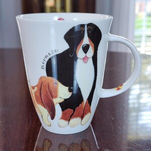 May include: A white ceramic mug featuring a colorful illustration of a Bernese Mountain Dog and a Basset Hound. The word Bernese is printed in cursive script next to the dogs. The mug has a curved handle and a smooth, glossy finish throughout.