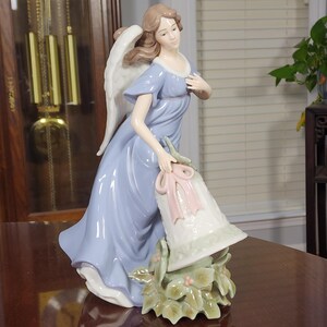 May include: A porcelain figurine of an angel with light brown hair and white wings, wearing a flowing periwinkle blue gown. The angel holds a white bell adorned with a soft pink ribbon, positioned above a base of green holly leaves and small berries.