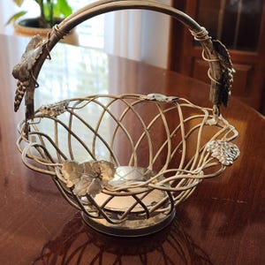 May include: A silver-toned metal basket with a handle, featuring a woven design. The basket is adorned with grape and leaf accents. The handle is arched, and the basket sits on a round base. The interior appears to be reflective.