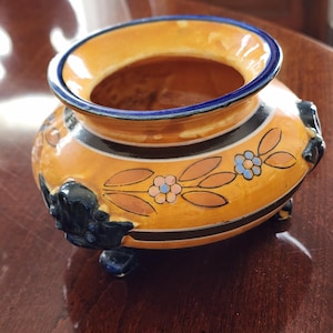 May include: A ceramic decorative bowl with a wide opening, featuring a vibrant orange base with black and blue accents. The bowl is adorned with floral and leaf designs in orange, blue, and pink. It has three small feet and decorative handles.