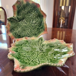 May include: Two leaf-shaped ceramic plates. The plates are green with detailed vein patterns and a reddish-orange trim. The plates are on a dark wooden surface, with a blurred background.