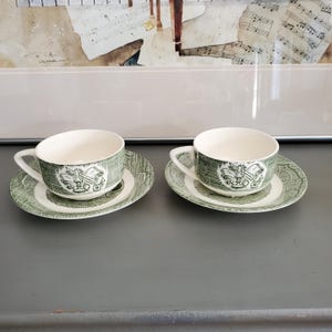 Może przedstawiać: Dwie pasujące filiżanki i spodki. Filiżanki i spodki są białe z zielonym wzorem. Filiżanki mają uchwyt i zielony pasek z wzorem. Spodki mają pasujący zielony wzór. W tle znajduje się oprawione dzieło sztuki.