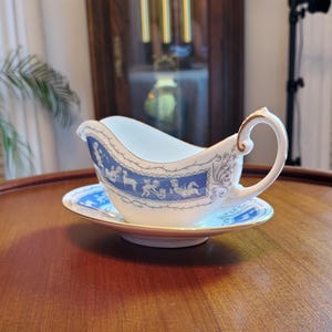 Könnte beinhalten: Eine weiße Porzellan-Sauciere mit passender Untertasse. Das Boot hat einen blau-weißen dekorativen Rand mit klassischen Figuren. Der Rand und der Griff sind mit goldenen Zierleisten versehen. Die Untertasse hat ein passendes blau-weißes Design.