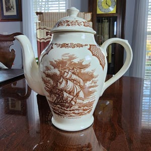 Puede incluir: Tetera de cerámica color crema con una ilustración marrón de un velero. La tetera tiene un pico curvo, un asa grande y una tapa con detalles florales. Las palabras "U.S.S. Constitution" están impresas en la base.