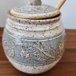 May include: A ceramic jar with a lid and a wooden spoon. The jar is light blue and white with a floral design. The lid has a knob on top. The jar is sitting on a wooden surface.