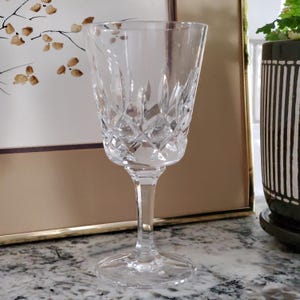 May include: A clear crystal wine glass with a diamond-cut pattern on the bowl. The glass has a long stem and a round base. The background includes a framed artwork and a decorative plant.
