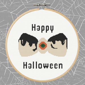 Puede incluir: Bordado de punto de cruz en un aro de madera con las palabras "Happy Halloween". El diseño presenta dos formas estilizadas en forma de huevo negro y beige con un pequeño centro naranja y verde. El fondo es un patrón de telaraña gris.