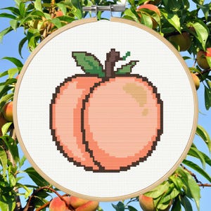 May include: A cross-stitch embroidery hoop with a pixelated peach design. The peach is in pink and orange with a brown outline, green leaves, and a brown stem. The embroidery is on white fabric, framed by a wooden hoop. The background shows a peach tree.