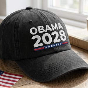 Könnte beinhalten: Schwarze Baseballkappe mit dem weißen Text "OBAMA 2028", mit einem rot-blauen Streifen und Sternenmuster. Die Kappe liegt auf einer Holzoberfläche, mit einer kleinen amerikanischen Flagge.