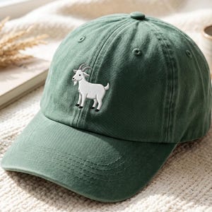 Op de afbeelding: Groene baseballcap met een gebogen klep en een kleine, witte geborduurde geit op de voorkant. De cap is gemaakt van een zachte stof en heeft een ronde bovenkant met een kleine knoop.