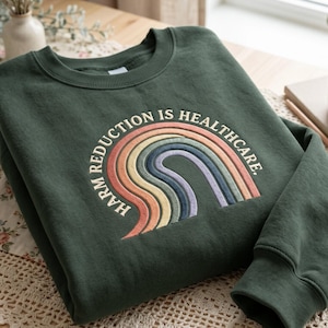 May include: A dark green sweatshirt with the embroidered text "Harm Reduction is Healthcare" above a rainbow design in pastel colors. The sweatshirt has a crew neck and long sleeves.