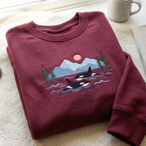 May include: Burgundy-colored sweatshirt with an embroidered design featuring two orcas swimming in water, mountains, trees, and a sun. The crew neck sweatshirt is made of a soft material.