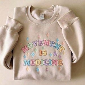 May include: A beige sweatshirt with the embroidered text "MOVEMENT IS MEDICINE" in pastel colors. The design includes stars and diamond shapes. The sweatshirt has a crew neck and ribbed cuffs.
