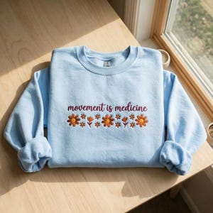 K&ouml;nnte beinhalten: Hellblaues Sweatshirt mit dem gestickten Text "movement is medicine" in Burgunderrot. Unter dem Text befinden sich gestickte orangefarbene und gelbe Blumen. Das Sweatshirt liegt auf einer Holzoberfl&auml;che.