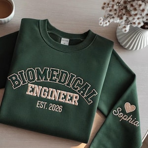 Op de afbeelding: Een donkergroene sweatshirt met de geborduurde tekst "BIOMEDICAL ENGINEER EST. 2026" in lichtbeige. De mouw heeft de naam "Sophia" en een hart. De sweatshirt is een casual kledingstuk.