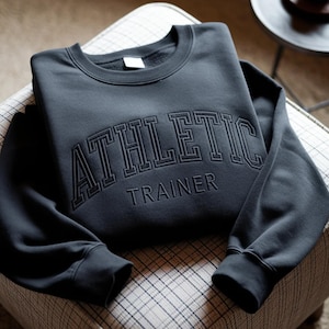 May include: A dark gray sweatshirt with the words "ATHLETIC TRAINER" embroidered across the chest. The sweatshirt is folded on a patterned chair, with the sleeves neatly arranged. The text is in a bold, block letter style.