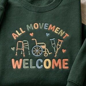 May include: Dark green sweatshirt with embroidered text and graphics. The text reads "ALL MOVEMENT WELCOME" in colorful letters. Graphics include a walker, wheelchair, and crutches, with heart accents.