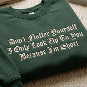 Pode incluir: Um moletom verde escuro com a frase bordada "Don't Flatter Yourself I Only Look Up To You Because I'm Short" em uma fonte decorativa. O moletom de gola redonda é feito de um material macio.