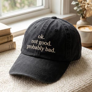 May include: A black baseball cap with a curved brim. The cap features the embroidered text "ok. not good. probably bad." The hat is made of a soft, worn fabric and has a button on top.