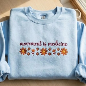 K&ouml;nnte beinhalten: Hellblaues Sweatshirt mit dem gestickten Schriftzug "movement is medicine" in Burgunderrot. Unter dem Text befindet sich eine Reihe von orangefarbenen und gelben gestickten Blumen. Das Sweatshirt hat lange &Auml;rmel.
