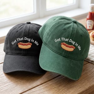 Puede incluir: Se muestran dos gorras de b&eacute;isbol, una negra y otra verde. Cada gorra presenta la frase bordada "Got That Dog In Me" sobre un dibujo de un perrito caliente. Las gorras est&aacute;n sobre una superficie de madera.
