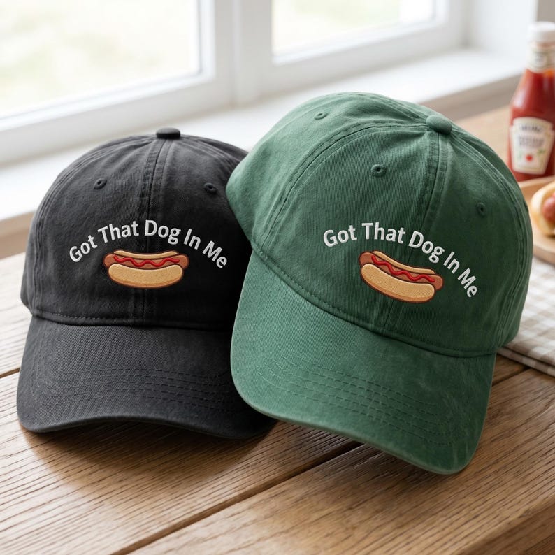 Puede incluir: Se muestran dos gorras de b&eacute;isbol, una negra y otra verde. Cada gorra presenta la frase bordada "Got That Dog In Me" sobre un dibujo de un perrito caliente. Las gorras est&aacute;n sobre una superficie de madera.