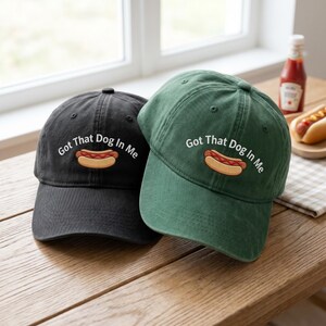 Puede incluir: Se muestran dos gorras de b&eacute;isbol, una negra y otra verde. Cada gorra tiene la frase "Got That Dog In Me" bordada sobre un dibujo de un perrito caliente. Las gorras est&aacute;n sobre una superficie de madera.