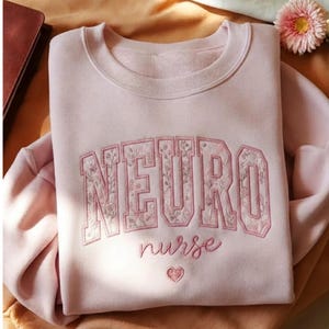 Puede incluir: Sudadera rosa claro con la palabra "NEURO" bordada en un diseño floral, con "nurse" debajo y un corazón. La sudadera de cuello redondo es de estilo casual.
