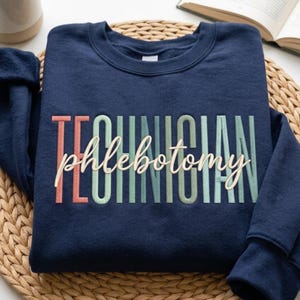 May include: Navy blue sweatshirt featuring the embroidered text "TECHNICIAN phlebotomy" in a mix of colours. The text is arranged vertically. The sweatshirt is displayed on a woven surface.