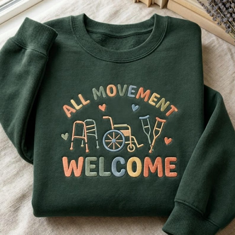 May include: Dark green sweatshirt with the embroidered text "ALL MOVEMENT WELCOME" in colorful letters. The design includes illustrations of a walker, wheelchair, and crutches, with small heart accents. The sweatshirt is made of a soft material.