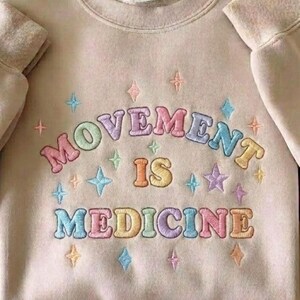 May include: Beige sweatshirt with the embroidered text "MOVEMENT IS MEDICINE" in pastel colors. The design includes star and diamond shapes in various colors. The sweatshirt has a crew neck and long sleeves.